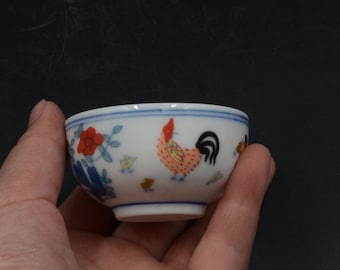 May include: Small white ceramic bowl with a hand-painted rooster and floral design. The rooster is red, orange, and yellow with a black tail. The bowl has a blue rim and is held in a hand.