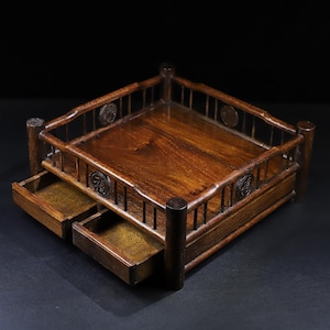 May include: A dark brown wooden display stand featuring a square top surface enclosed by a railing with vertical slats. The stand includes two small pull-out drawers at the front, each adorned with a circular emblem containing a Chinese character.