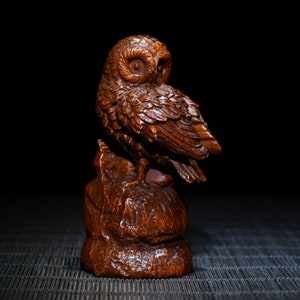 Puede incluir: Escultura de madera detallada de un búho, posada sobre una base texturizada. El búho está tallado con intrincados detalles de plumas y tiene una mirada enfocada. La escultura es de color marrón cálido, sobre un fondo oscuro.