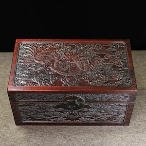 Pode incluir: Uma caixa de madeira vermelha escura com entalhes intrincados de um dragão e nuvens. A caixa tem uma tampa articulada e uma trava de metal.