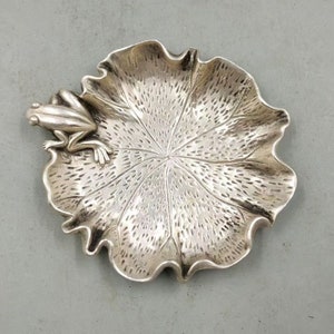 Può includere: Un piatto decorativo color argento a forma di ninfea, con una rana appollaiata sul bordo. Il piatto ha una superficie strutturata e venature delle foglie dettagliate. La rana è anch'essa argentata e presenta dettagli intricati.