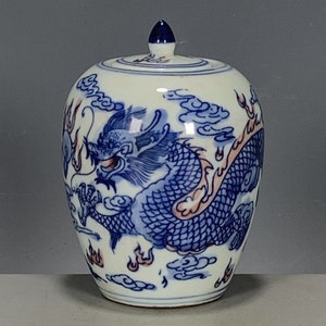 Puede incluir: Un jarrón de porcelana blanca con un diseño de dragón azul y rojo. El jarrón tiene una tapa y está decorado con nubes y llamas.