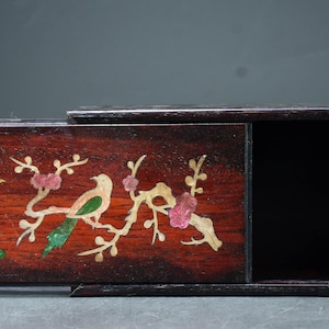 May include: A rectangular wooden box with a sliding lid. The lid features inlaid birds, flowers, and branches in gold, green, and pink against a reddish-brown background. The box is dark brown.