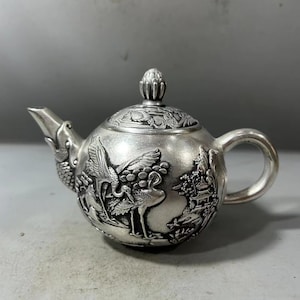 Puede incluir: Una tetera de plata con un diseño intrincado de grullas y una pagoda. La tetera tiene un cuerpo redondo y un pequeño pico. El asa es curva y la tapa tiene un pequeño pomo.