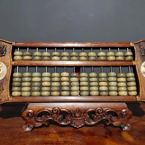 Pode incluir: Um ábaco de madeira vintage com duas fileiras de contas verdes. A moldura é marrom escuro com detalhes esculpidos e caracteres chineses. O ábaco está sobre um suporte de madeira decorativo.