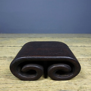 May include: A dark brown wooden scroll stand with a smooth, polished finish. The stand is shaped like a rolled scroll and has two rounded ends.