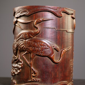 Puede incluir: Un recipiente de bambú marrón oscuro con intrincados tallados de grullas y nubes. El recipiente es cilíndrico y tiene un acabado liso y pulido.