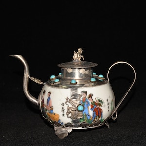 Puede incluir: Una tetera de porcelana blanca con asa, boquilla y tapa plateadas. La tapa está adornada con una pequeña escultura plateada y piedras turquesas. El cuerpo presenta una escena pintada de figuras con vestimenta tradicional. La tetera descansa sobre tres patas plateadas.