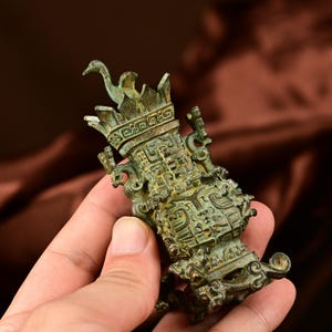 May include: A detailed bronze-colored decorative vessel with intricate carvings and a bird-like figure on top. The vessel has a weathered appearance, suggesting age, and is held in a hand against a blurred background.