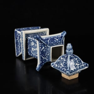Puede incluir: Un recipiente de porcelana azul y blanco con forma cuadrada y tapa a juego. El recipiente presenta intrincados diseños florales y geométricos. La tapa está a un lado, revelando el interior del recipiente. La pieza está sobre un fondo negro.