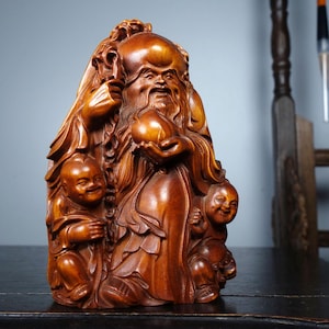 Puede incluir: Escultura de madera detallada que representa a un hombre barbudo sosteniendo una fruta, rodeado de tres niños sonrientes. La escultura está tallada en madera de color marrón rojizo cálido, con detalles intrincados en la ropa y los rasgos faciales. Las figuras se encuentran sobre un fondo neutro.