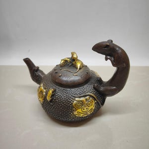 Puede incluir: Una tetera marrón con un asa en forma de rata y una tapa con dos ratas en la parte superior. La tetera está decorada con detalles dorados y tiene una superficie texturizada.
