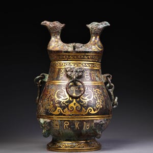 May include: A bronze vessel with a dark patina and gold inlay featuring two bird-shaped finials on the lid. The body displays intricate gold scrollwork, animal motifs, and ancient characters. It includes side handles with rings and a base.