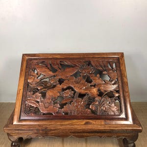 May include: A rectangular wooden display stand with detailed carvings. The stand features a relief of aquatic plants and fish. The wood has a rich, reddish-brown hue, and the stand rests on four rounded feet. The carved panel is framed.