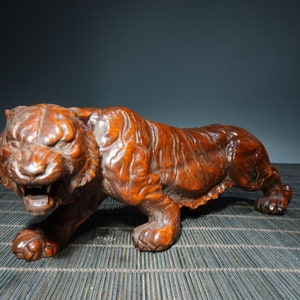 Op de afbeelding: Gedetailleerde houten sculptuur van een tijger in een hurkende positie. De tijger is gesneden met ingewikkelde details, die zijn vacht, gezichtskenmerken en gespierde bouw laten zien. De sculptuur heeft een warme bruine kleur.