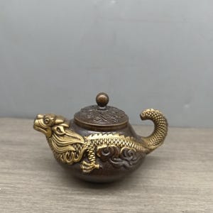 Puede incluir: Una tetera pequeña de bronce con un diseño de dragón. La tetera tiene un dragón dorado en el lateral y un pomo dorado en la tapa.