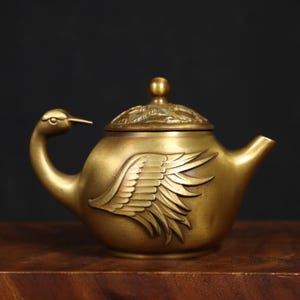 Può includere: Una teiera color ottone con un manico a forma di cigno e un design ad ala sul lato. Il coperchio è adornato con un motivo decorativo e un piccolo pomello. La teiera è in metallo e ha un beccuccio.