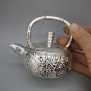 Puede incluir: Una tetera de plata con un asa de bambú y un diseño detallado de hojas y ramas de bambú. La tetera tiene una tapa y un pico.