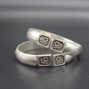 May include: Two silver bangles with a floral design. Each bangle features a rectangular clasp with a detailed flower pattern. The bracelets are displayed on a dark gray surface, showcasing their polished finish and intricate details.