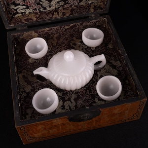 Könnte beinhalten: Ein weißes Jade-Tee-Set in einer Holzkiste mit brauner und goldener Musterung. Das Set enthält eine Teekanne und vier Tassen.