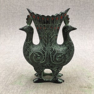 Op de afbeelding: Een groene en zwarte metalen vaas met twee pauwen-vormige handvatten. De vaas is versierd met ingewikkelde bloemmotieven en rode accenten.