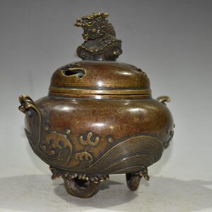 May include: A bronze incense burner with a lid and a dragon figure. The burner has a rounded body with wave patterns and small handles. It stands on three curved feet. The overall colour is bronze.