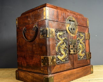 Op de afbeelding: Een houten juwelendoos met messing accenten en drakengravures. De doos heeft twee deuren met een messing sluiting en een handvat aan de zijkant.