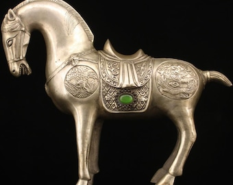 May include: A silver horse figurine with intricate details, including a saddle with a green gemstone. The horse is standing on a black background.