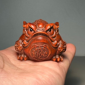 Könnte beinhalten: Eine detaillierte, rotbraune Holzschnitzerei einer froschartigen Kreatur mit wütendem Gesichtsausdruck. Die Skulptur weist komplizierte Details auf, darunter strukturierte Haut, hervorstehende Augen und ein dekoratives Emblem auf der Brust. Die Schnitzerei wird in einer Hand gehalten.