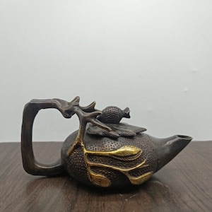 May include: A small, bronze teapot with a detailed design of a tree branch and leaves. The teapot has a gold-coloured finish on the leaves and a small, round knob on the lid.