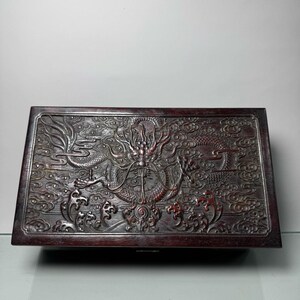 Puede incluir: Una caja de madera oscura con un diseño de dragón tallado en la tapa. El dragón es de color plateado y está rodeado de nubes.