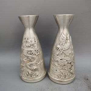 Puede incluir: Un par de jarrones de plata con intrincados diseños en relieve. Un jarrón presenta un dragón, mientras que el otro presenta un fénix.