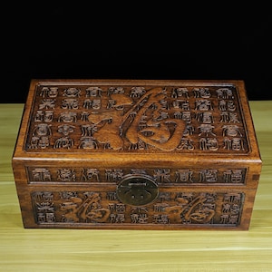Może przedstawiać: Drewniana skrzynia w ciemnym odcieniu brązu, z misternymi rzeźbami i metalowym zamkiem. Skrzynia jest ozdobiona chińskimi znakami.