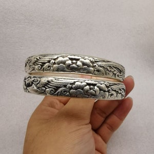 May include: Two silver bracelets with intricate floral and cloud designs. The bracelets are stacked on top of each other and have a polished finish.