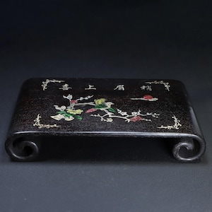 May include: Dark brown wooden display stand with a curved base and decorative floral and bird inlay. The stand features a rectangular top with a Chinese inscription and ornate corner designs. The bird is perched on a flowering branch.