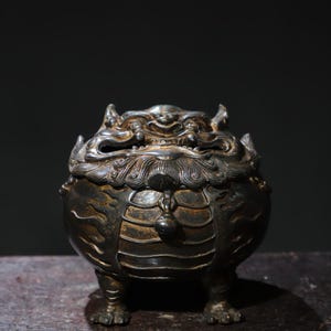 May include: A dark bronze incense burner shaped like a mythical creature with a fierce expression. The burner has intricate details and a textured surface.