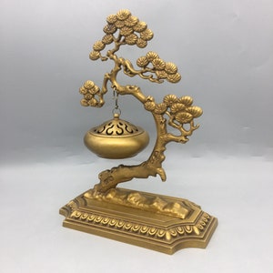 May include: A gold metal incense burner shaped like a pine tree with a hanging bowl. The bowl has a decorative design and is suspended from the tree by a chain.