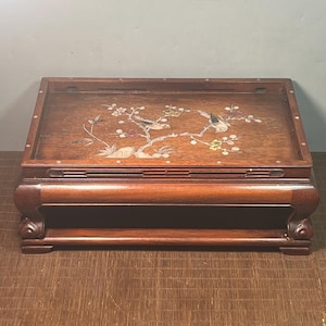 May include: A rectangular dark brown wooden display stand featuring a flat top surface with an inlaid mother-of-pearl design of two birds perched on flowering branches. The stand has a raised border with small circular inlays and curved legs.