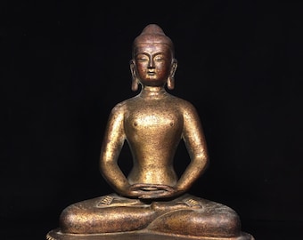 Op de afbeelding: Een zittend figuur in een meditatieve houding met de handen in de schoot. Het beeld heeft een metallic goud- en bronskleurige afwerking met een verweerde textuur. Het figuur rust op een voetstuk versierd met een uitgesneden lotusbloempatroon.