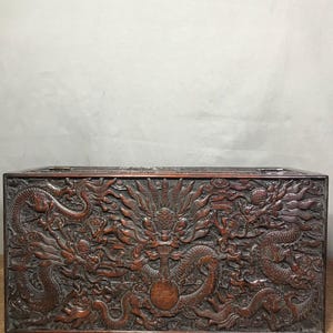 Pode incluir: Um baú de madeira escura com entalhes intrincados de dragões e um orbe central. O baú está fechado e tem uma forma retangular.