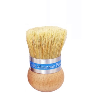 May include: A round wooden brush with light tan natural bristles and a silver metal ferrule. A blue label is wrapped around the ferrule with the white text Chalk Mountain Brush printed on it. The handle is a smooth, rounded, light-colored wood.