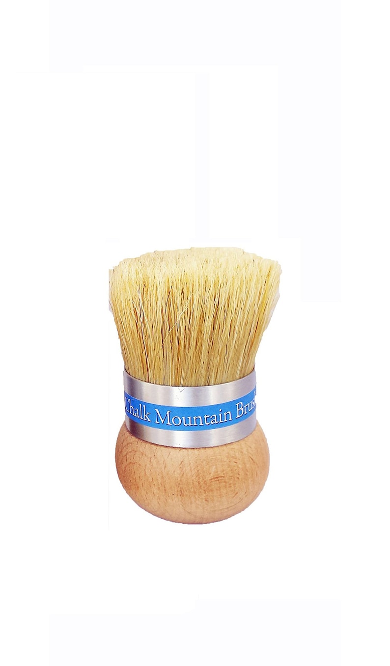 May include: A round wooden brush with light tan natural bristles and a silver metal ferrule. A blue label is wrapped around the ferrule with the white text Chalk Mountain Brush printed on it. The handle is a smooth, rounded, light-colored wood.