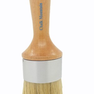 May include: A round paint brush with a light wood handle, a blue painted top, and a silver metal ferrule. The text Chalk Mountain is printed in blue on the wooden handle. The brush features long, light-colored natural bristles at the base.