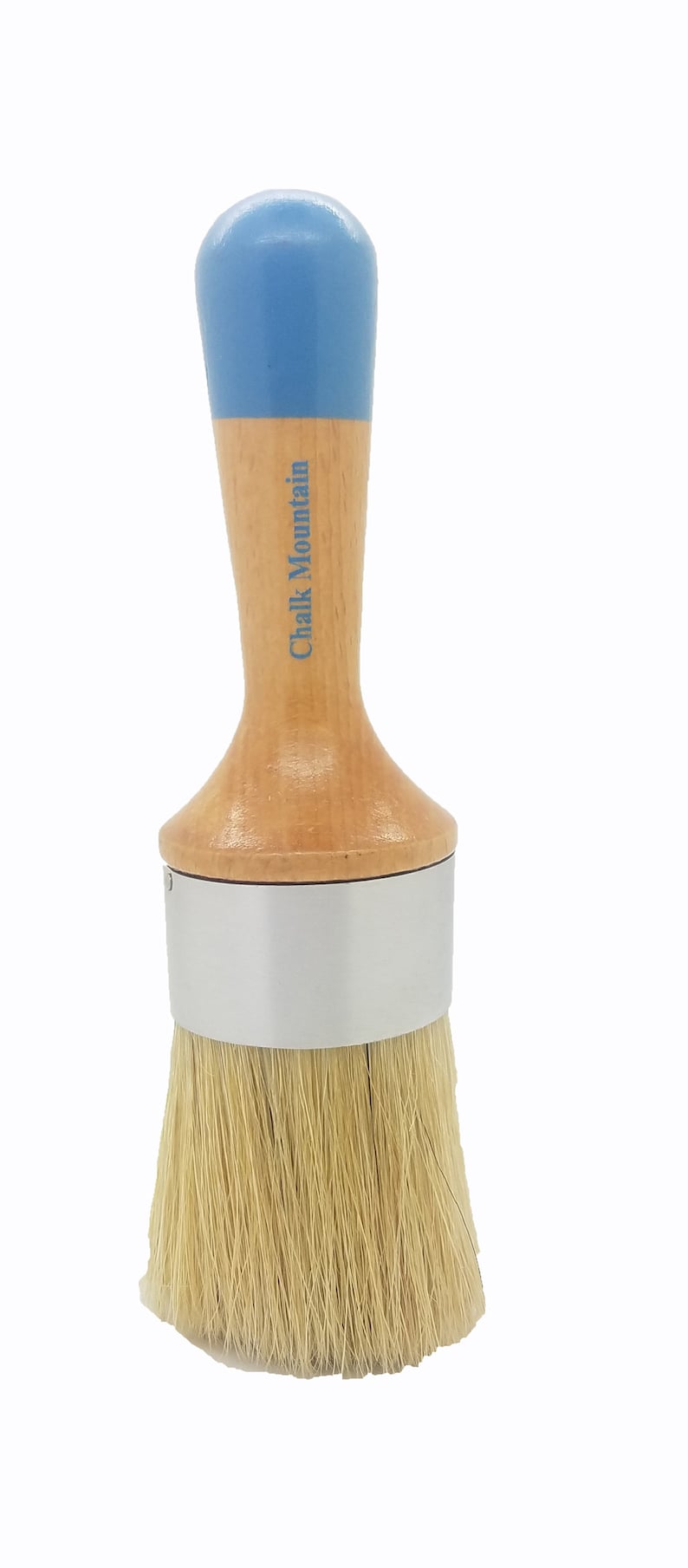 May include: A round paint brush with a light wood handle, a blue painted top, and a silver metal ferrule. The text Chalk Mountain is printed in blue on the wooden handle. The brush features long, light-colored natural bristles at the base.