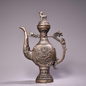 May include: Silver-colored decorative teapot with intricate dragon and animal designs. The teapot features a long spout, a handle with a dragon head, and a lid with a small animal figure. The body of the teapot is adorned with a detailed dragon relief.