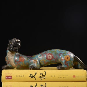 May include: A detailed, antique-style sculpture of a mythical creature, possibly a dragon or lion, with a turquoise body adorned with floral patterns in red, orange, and yellow. The creature rests on yellow books against a black background.