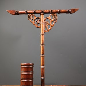 May include: A dark brown wooden calligraphy brush holder with a bamboo-style design. The holder features a horizontal bar with pointed ends, a central support, and a small cylindrical cup. The base is rectangular.