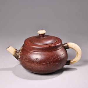 Könnte beinhalten: Eine braune Keramik-Teekanne mit gebogenem Griff und Ausguss, beide mit cremefarbenen Akzenten. Der Deckel hat einen kleinen, runden, cremefarbenen Knopf. Auf dem Körper der Teekanne sind florale Designs eingeätzt.