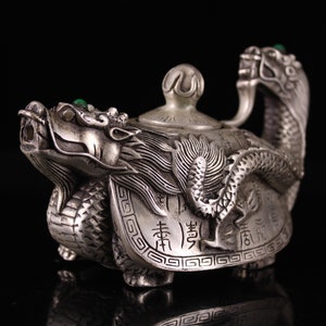 Puede incluir: Una tetera de plata con forma de dragón con detalles de piedras preciosas verdes. La tetera tiene detalles intrincados y caracteres chinos en el cuerpo.