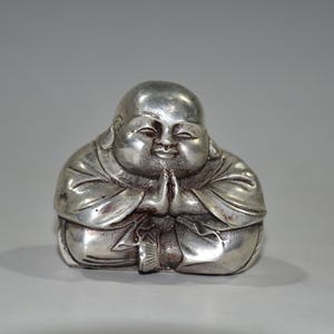 Puede incluir: Una estatua de Buda plateada en posición sentada con las manos juntas en oración. La figura tiene una barriga redonda y una expresión serena. La estatua está hecha de un material metálico brillante.